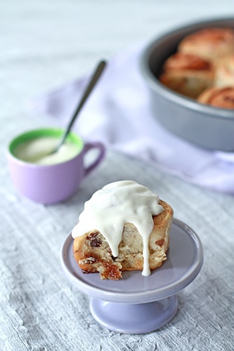 Cinnamon raisin bun with cream cheese frosting on a mini purple cake stand with frosting and pan of buns in background