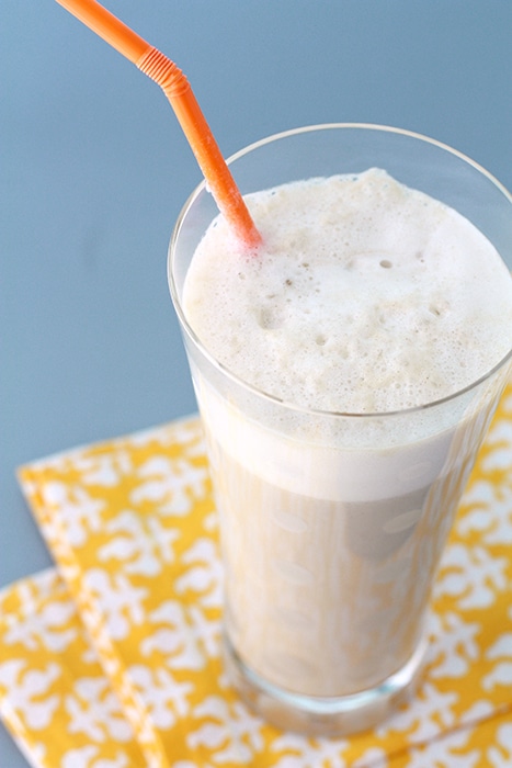 Coffee banana smoothie in a tall clear glass with orange straw, foamy on top