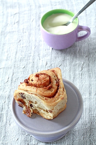 Cinnamon raisin bun on miniature purple cake stand, unglazed with icing in background ready to be spread on the bun