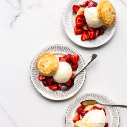 Three ice cream strawberry shortcakes on small dessert plates on white marble surface