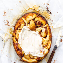 Cinnamon buns on parchment paper being frosted with cream cheese frosting using an offset spatula